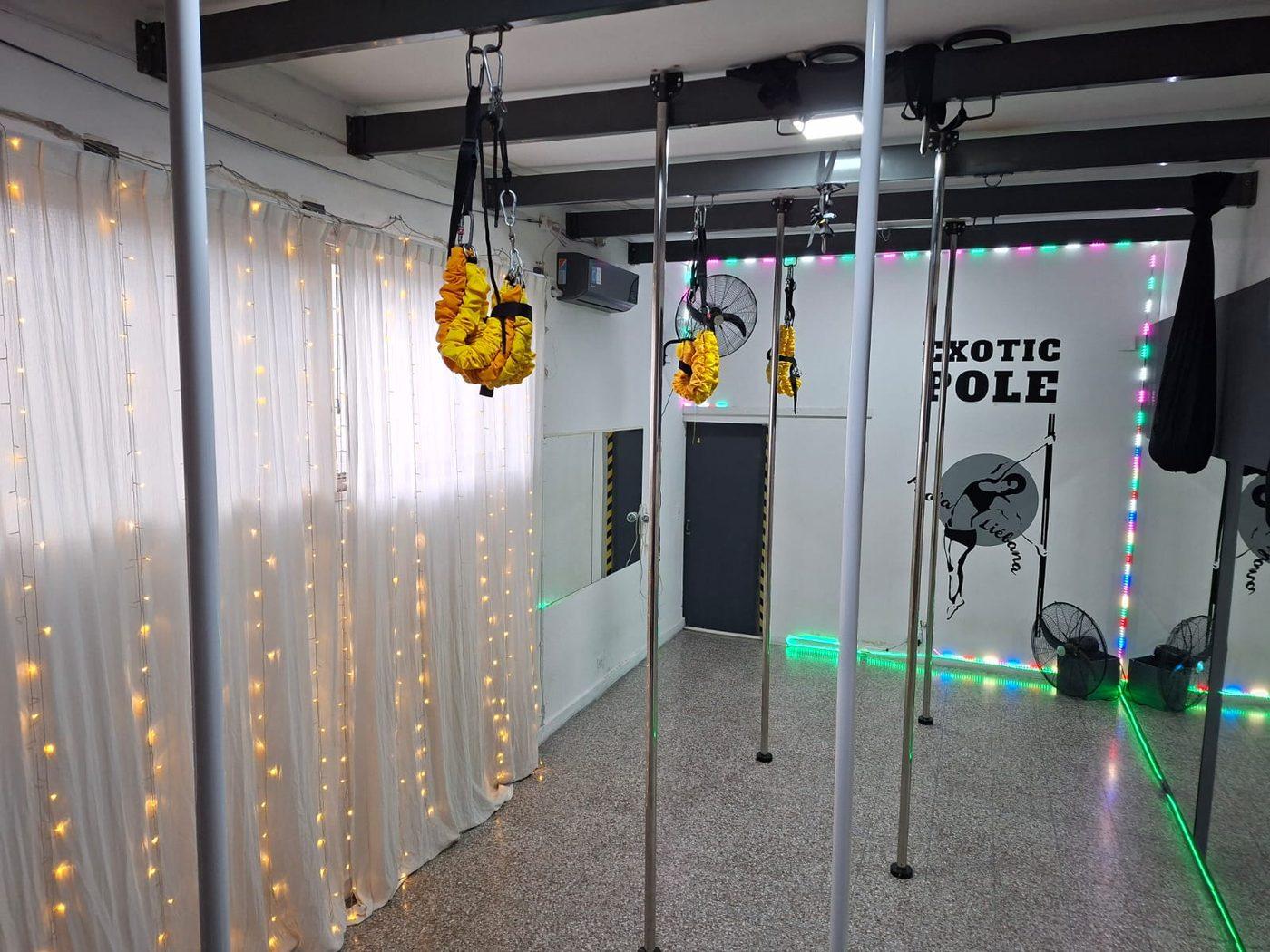 Sala de la sede Pacheco con los caños montados, arneses de bungee amarillos colgando de las vigas, cortina de luces y el mural EXOTIC POLE.