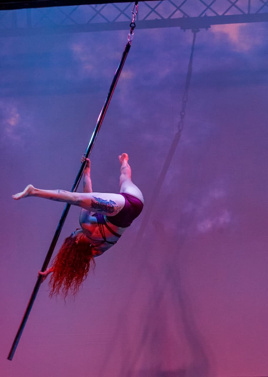 Paola Liébana suspendida en el caño de pole flying, que cuelga de una estructura con cadenas, en un escenario con luces violetas.
